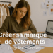 Entrepreneuse assise à une table avec des vêtements et un ordinateur portable affichant un logo, travaillant sur des idées pour créer sa marque de vêtements.